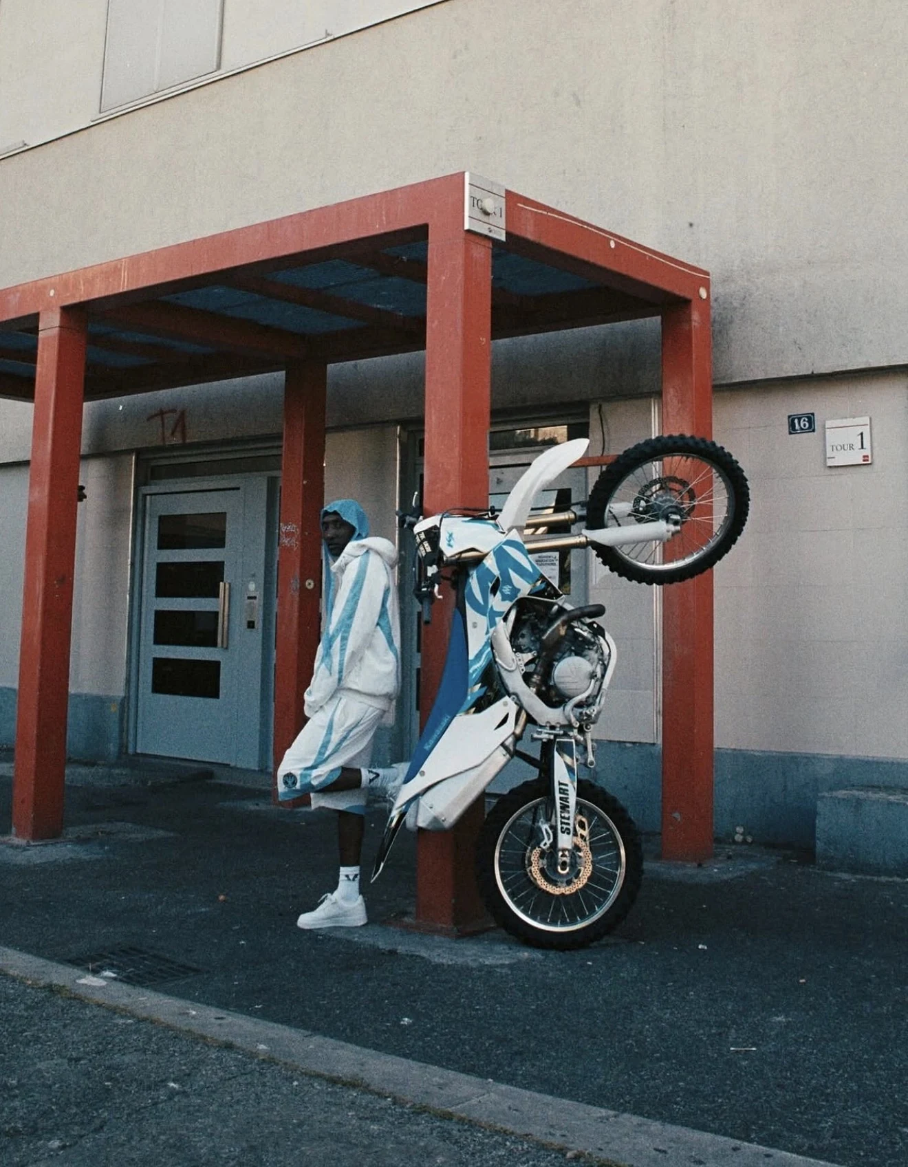Model standing beside a blue and white dirt bike under a red frame structure
